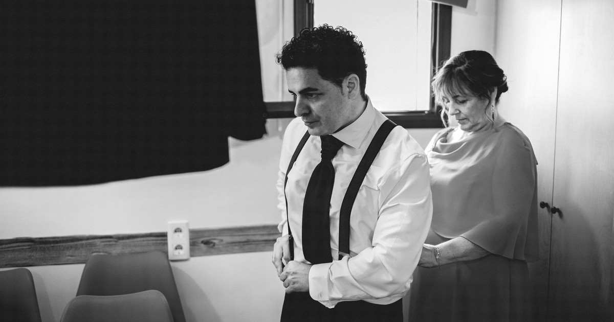 Black and white photo of a man in formal wear with a woman assisting him indoors.
