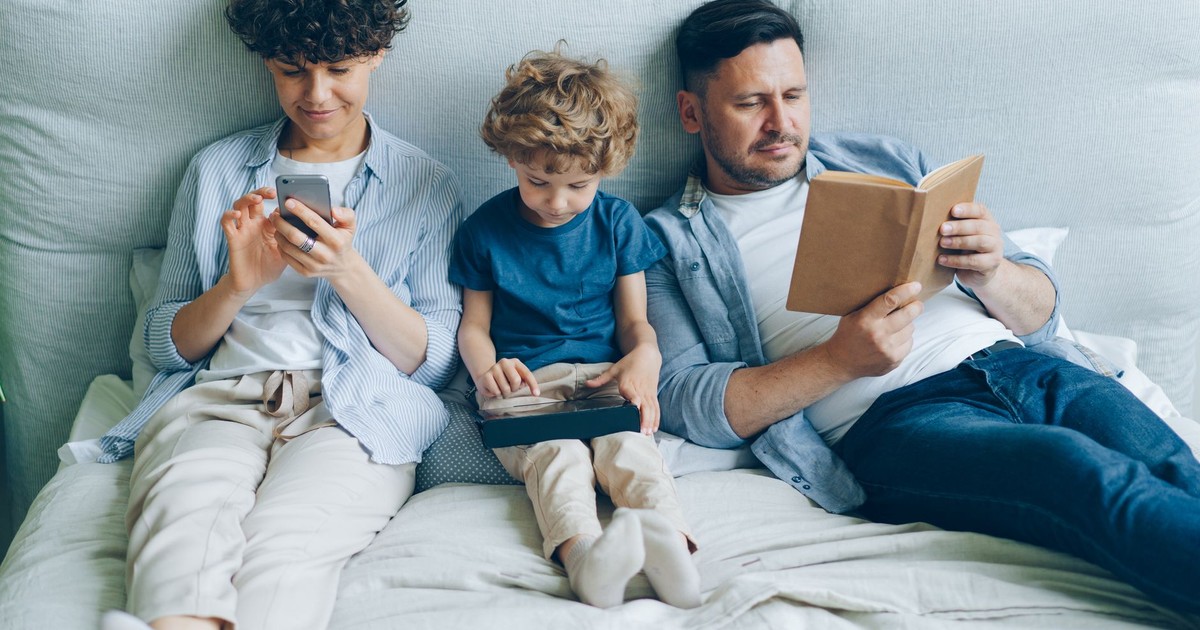 A family relaxing on bed with a smartphone, tablet, and book, symbolizing modern family life.