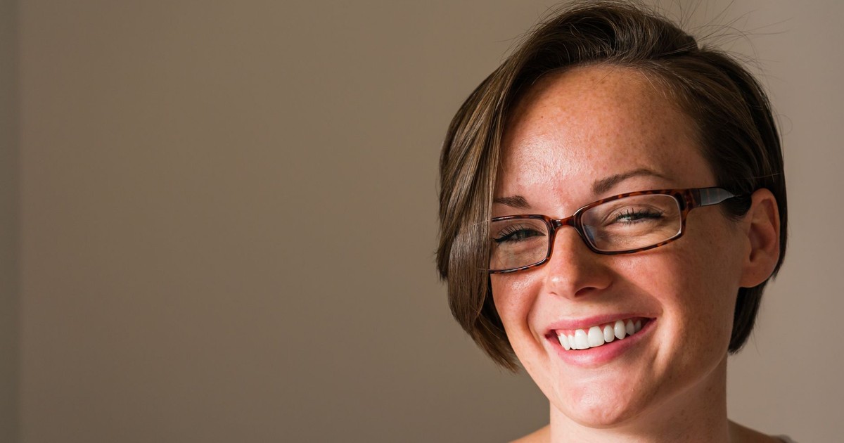 A cheerful woman with short hair and glasses smiles warmly indoors, exuding positivity.