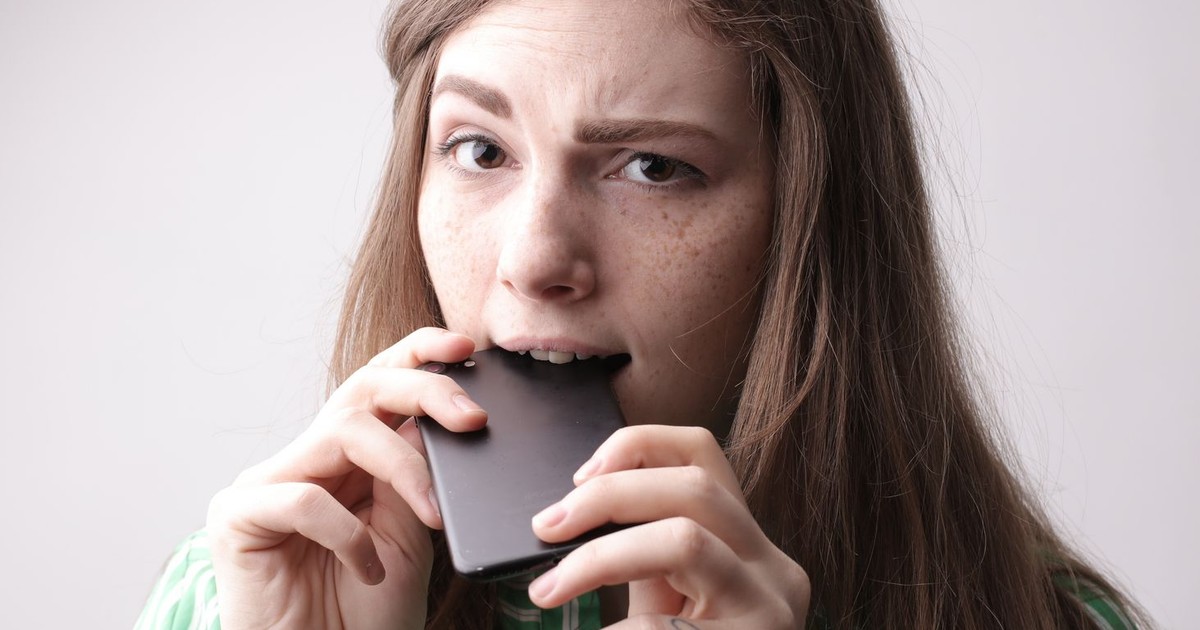Nervous young female in casual clothing standing with cellphone in mouth after unpleasant phone call on gray background in studio