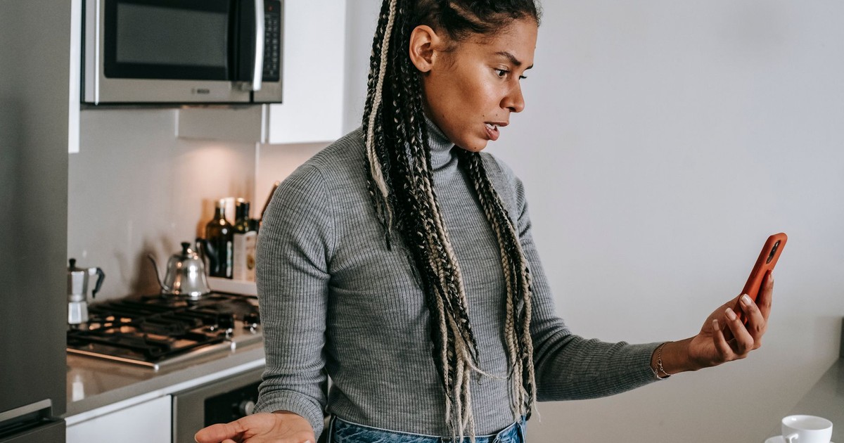 Serious young black lady with Afro braids in casual clothes gesticulating while having unpleasant conversation via video chat on smartphone in modern kitchen