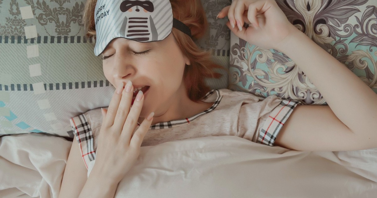Top view of female with sleep mask wearing pajama covering mouth with hand while lying on bed with closed eyes under blanket during bedtime