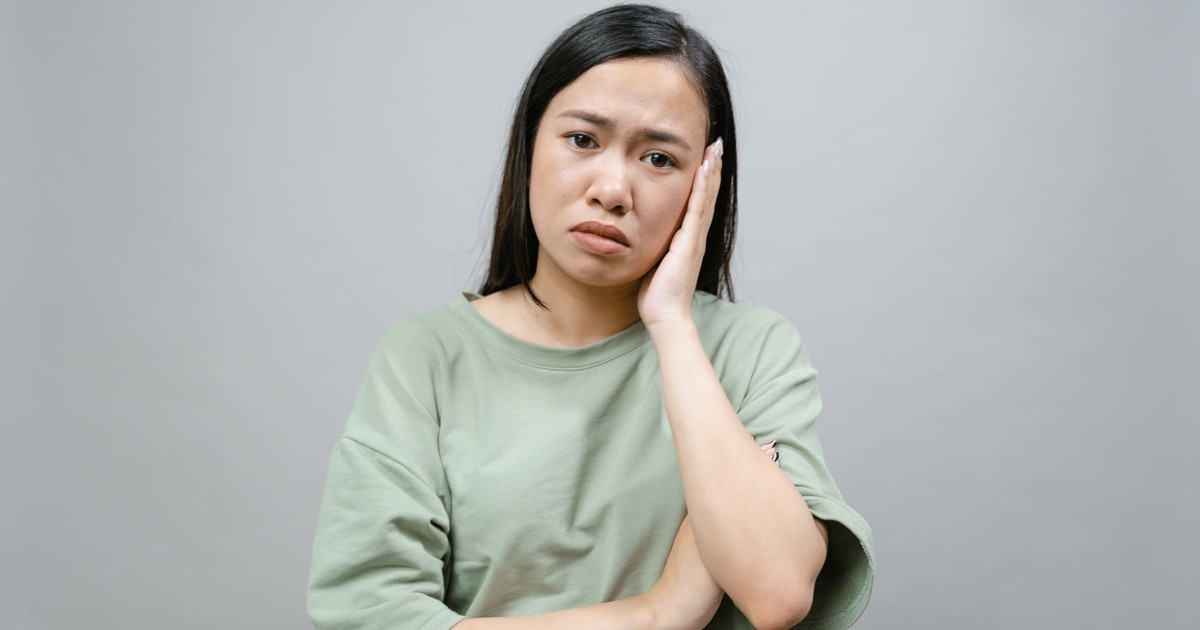 Portrait of a pensive woman indoors with a hand on her face expressing concern, on a grey background.