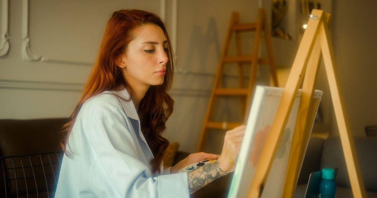 A focused young woman artist with red hair painting on a canvas in an art studio.