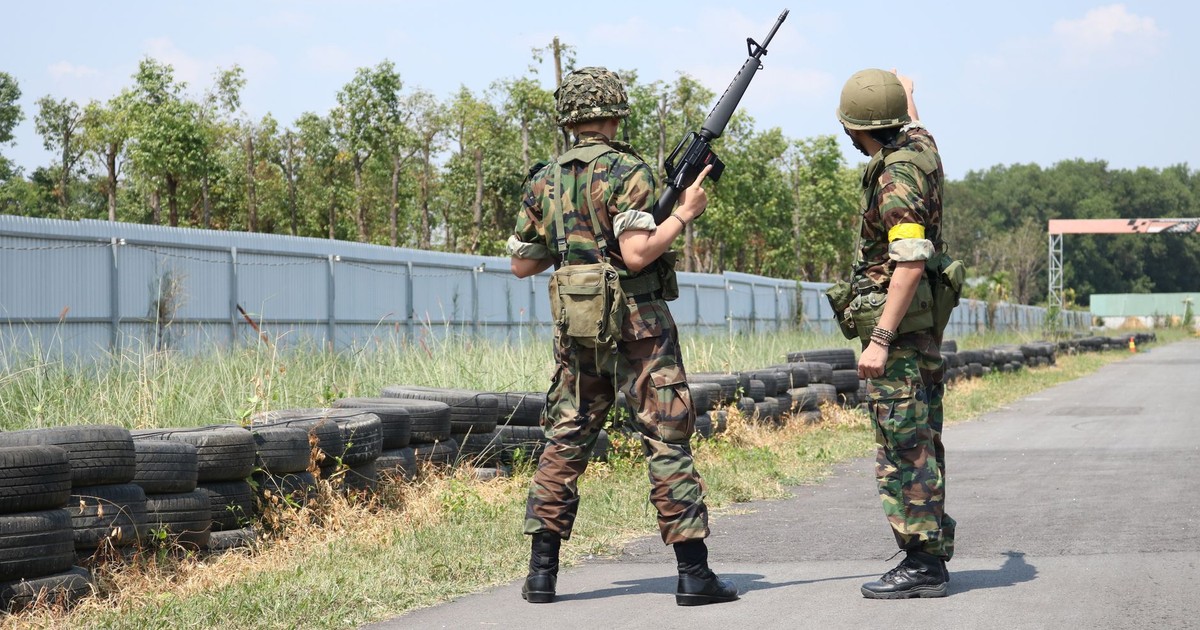 Two soldiers in camouflage uniforms with weapons stand outside on a training field.