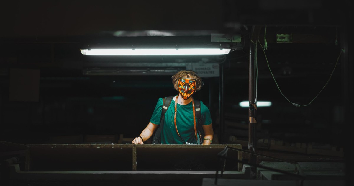 A masked person stands under a fluorescent light in an industrial environment, creating a mysterious and moody atmosphere.