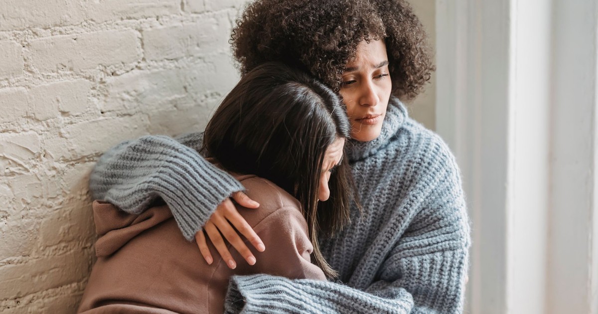 Unhappy African American woman in casual wear hugging and consoling female friend while sitting near wall in room at home