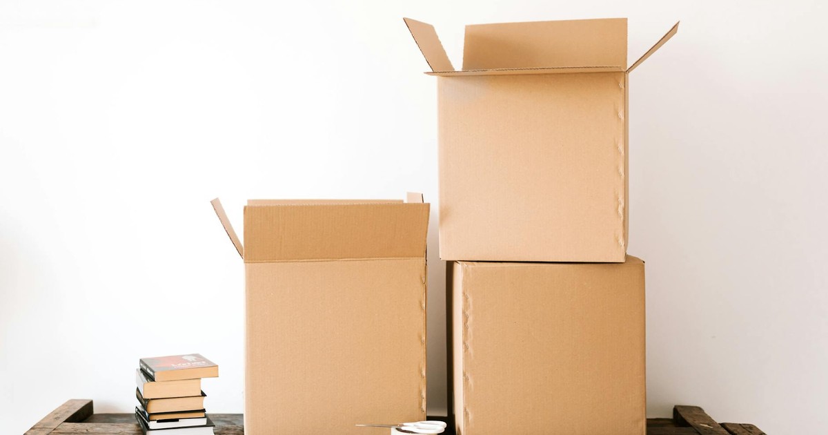 Opened cardboard boxes and stack of books placed on wooden shabby table near tape with scissors against white plain wall