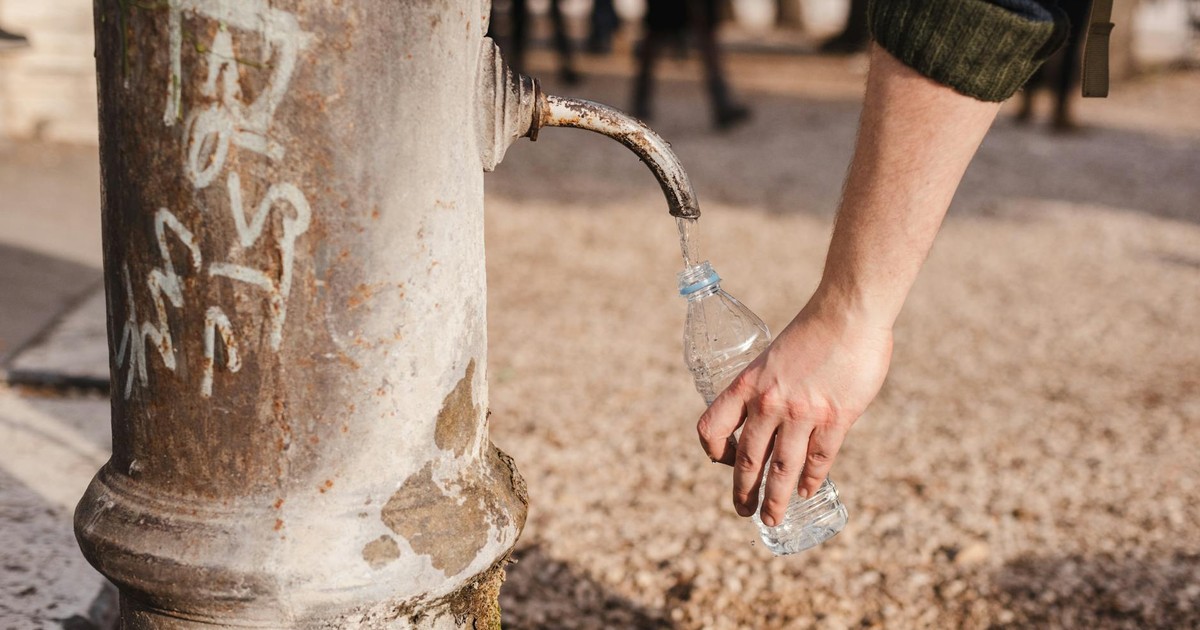 Crop unrecognizable person with bottle under old fountain tap with aqua flow on walkway in sunny town
