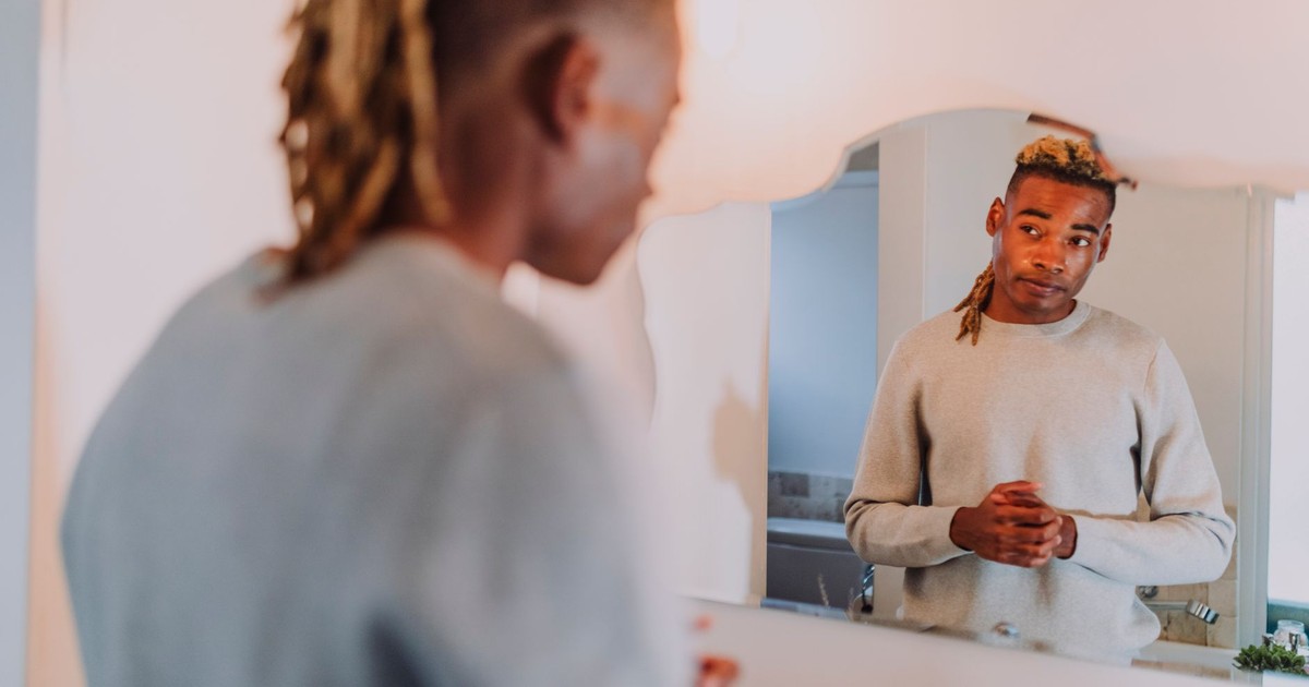 A man with dreadlocks stands contemplatively before a bathroom mirror.