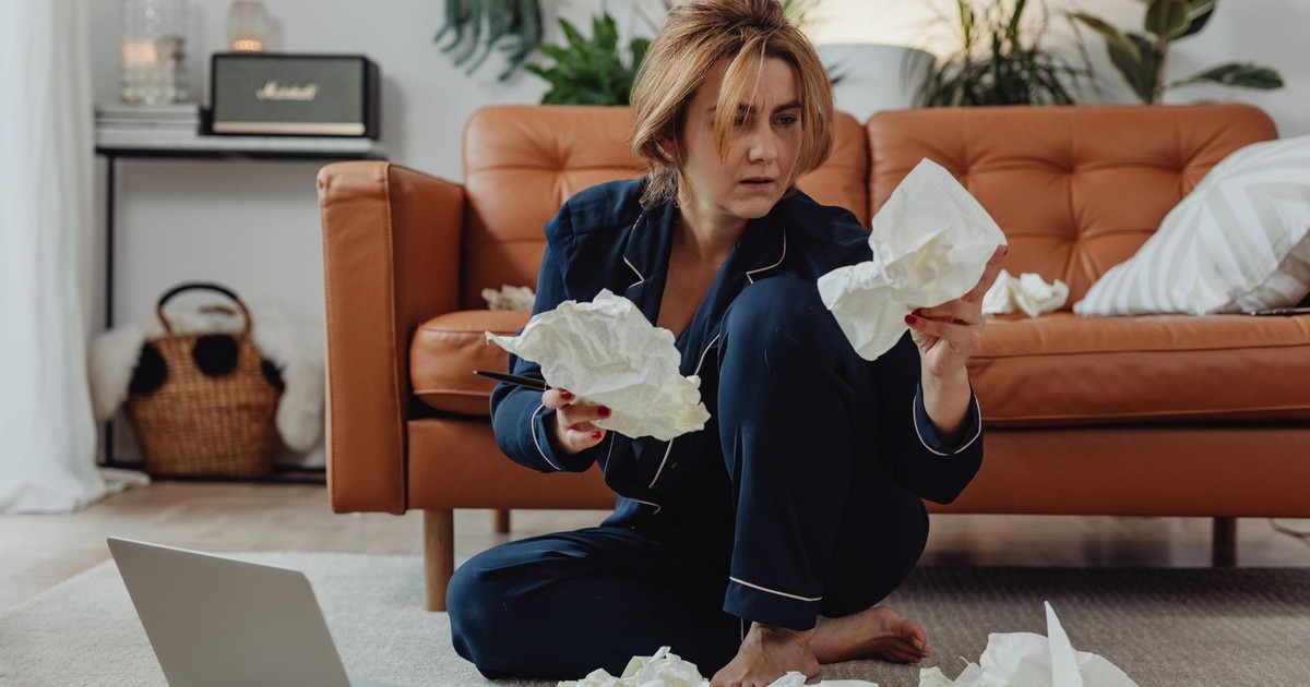 A woman in comfortable loungewear surrounded by crumpled papers, working remotely from home.