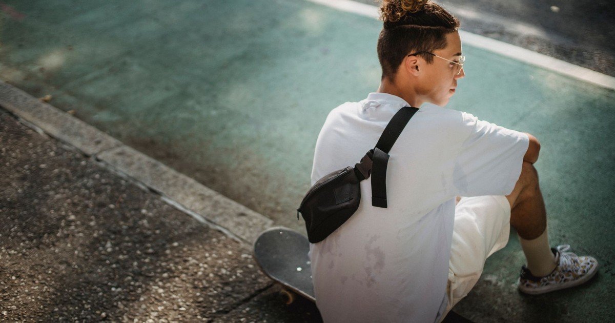 Back view of young ethnic male millennial with stylish hairstyle in white t shirt and shorts chilling on street sitting on skateboard after training