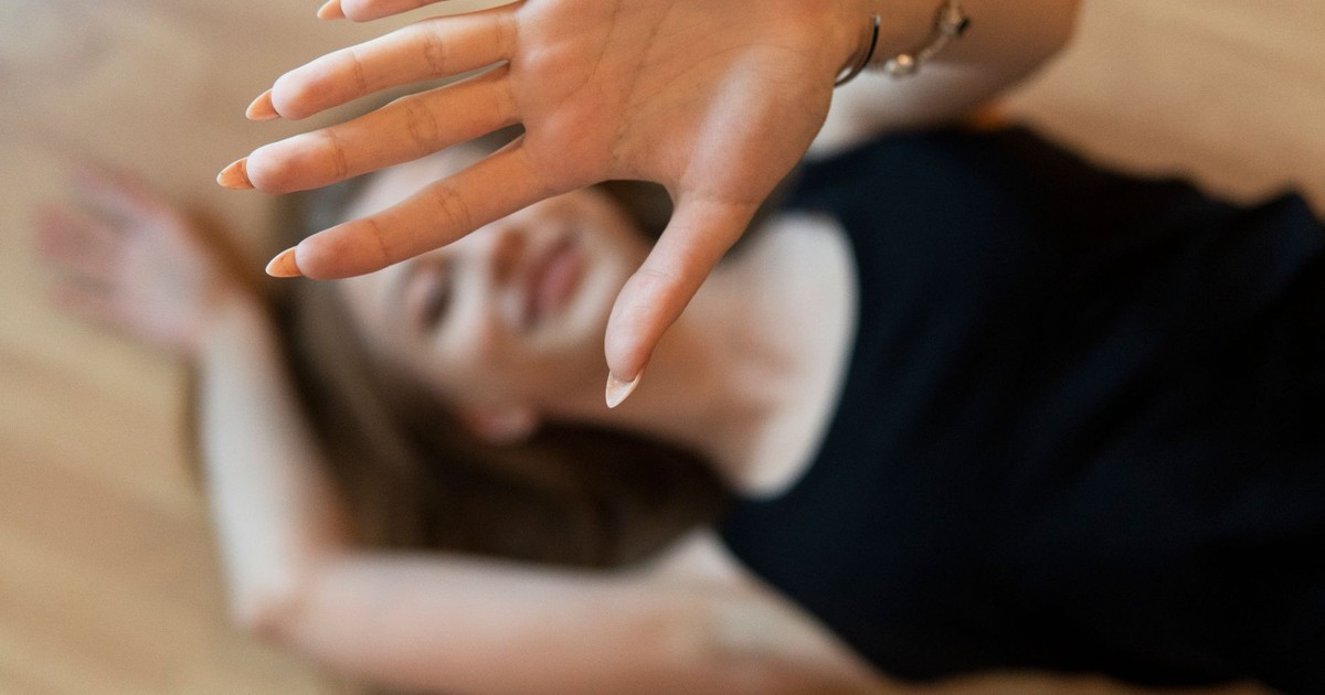 A woman lying on a wooden floor with a hand gesture blocking the frame, smiling naturally.