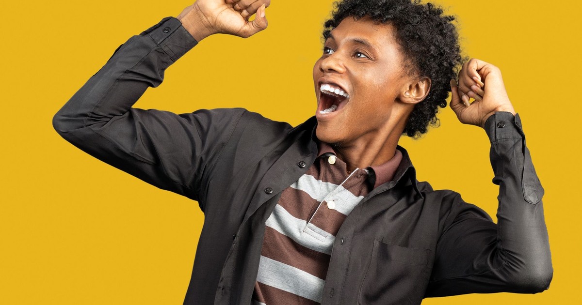 Expressive African American male wearing casual clothes standing against yellow background in studio and raising fists in triumph while looking away and screaming happily