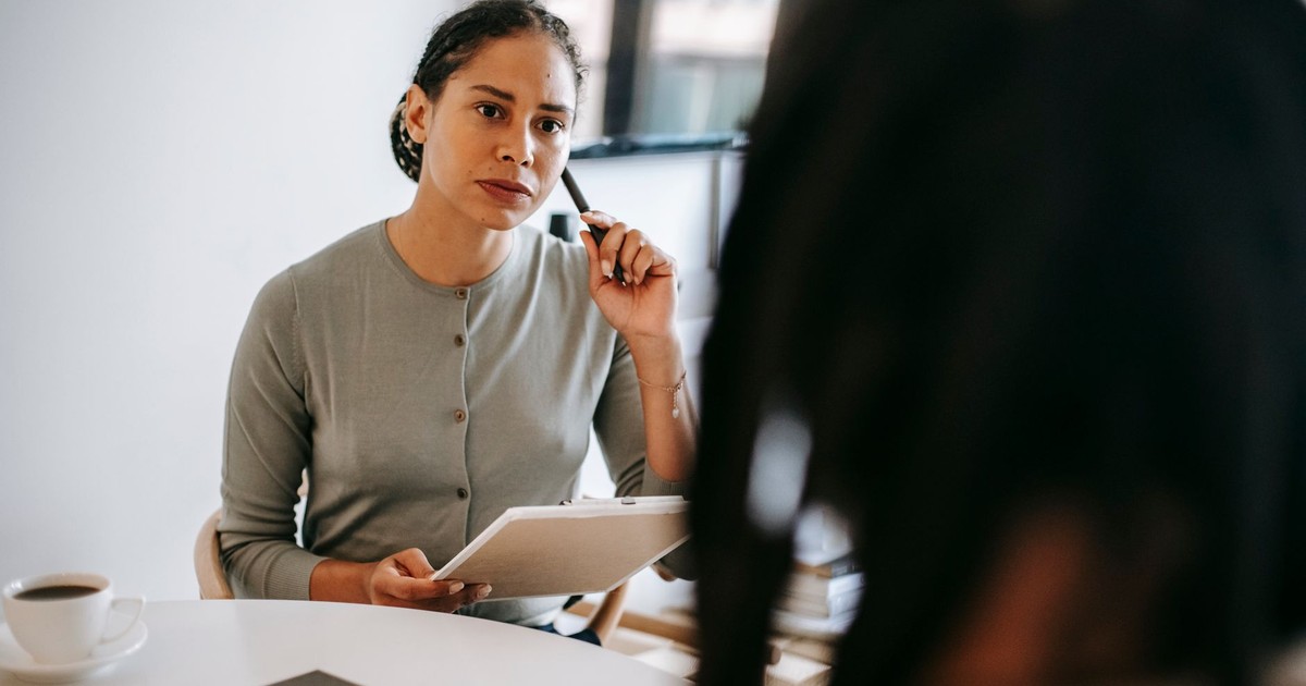 Focused ethnic female counselor in formal wear with clipboard and pen listening to patient problems during psychotherapy session