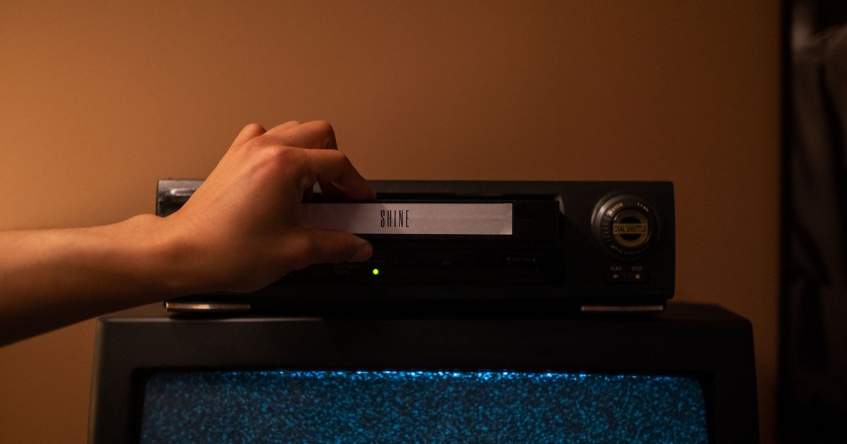 A hand inserting a VHS tape labeled 'SHINE' into a vintage player. Nostalgic home movie setup.