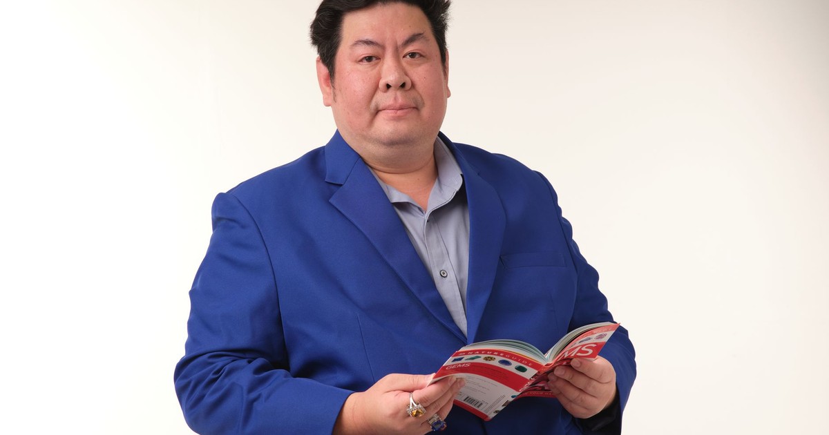 Confident businessman in a blue suit reading a magazine, sitting against a plain background.