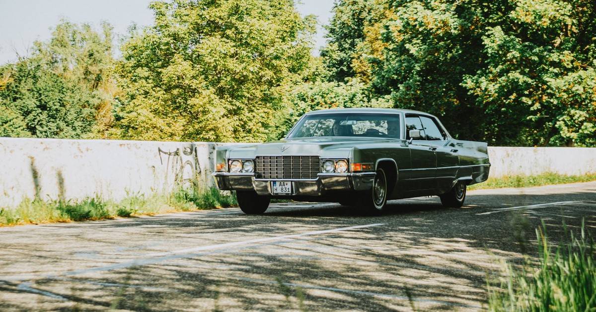 Classic green sedan from 1970s driving on a sunny road surrounded by trees.