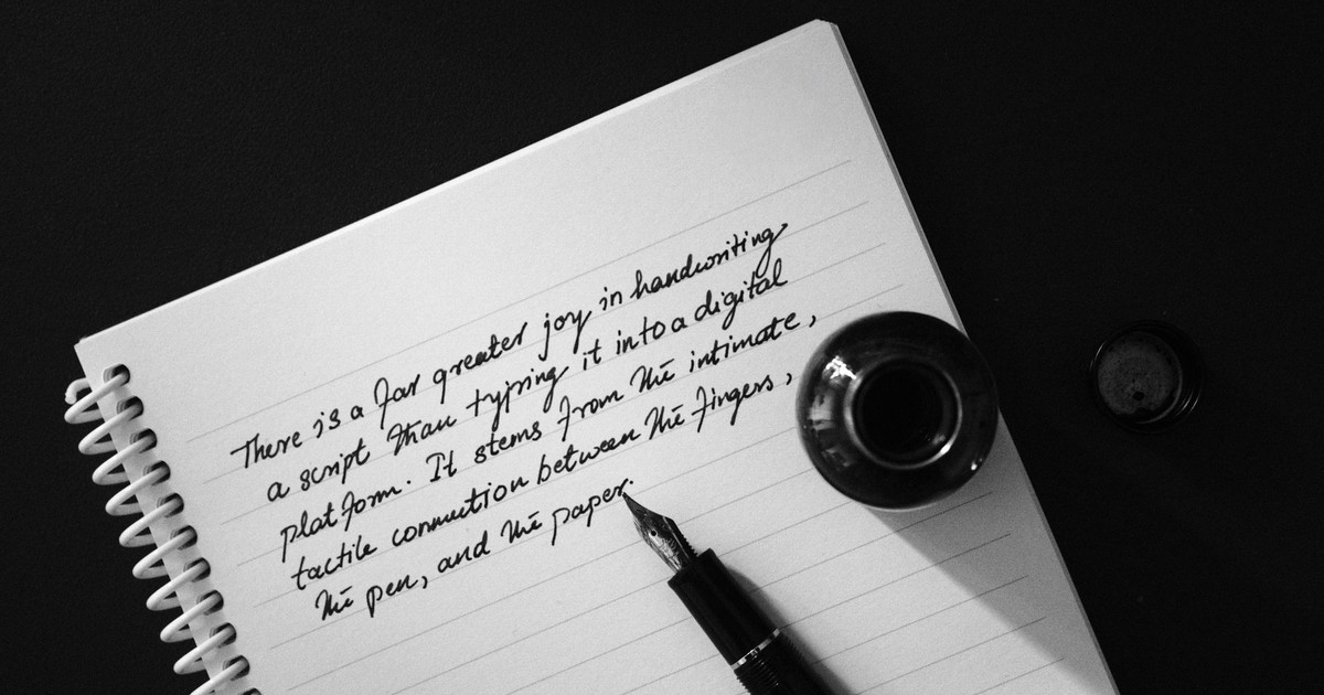 Black and white image of handwritten script with a fountain pen and ink bottle on a notebook.