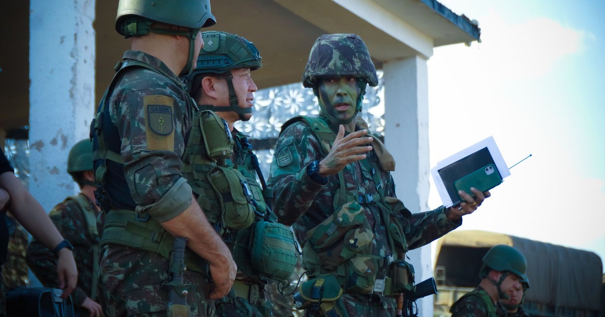 Group of soldiers in camouflage attending a briefing outdoors with tactical gear and helmets.