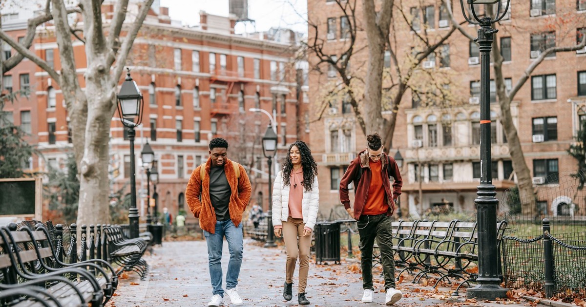 Full length of young positive multiracial friends in casual clothes walking in city park after studies on autumn day