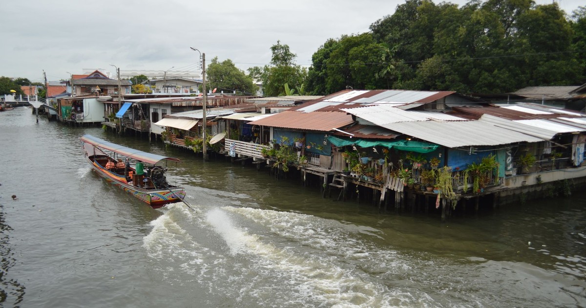 A scenic view of a traditional wooden boat navigating a canal beside rustic waterfront houses in Thailand.