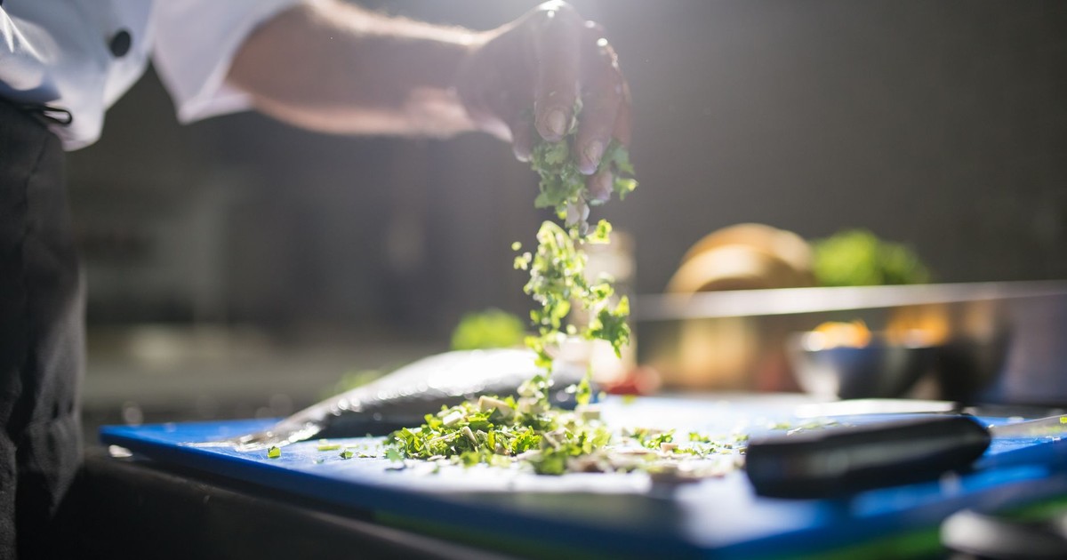 Chef's hand preparing chopped green vegetables on a cutting board in a kitchen.