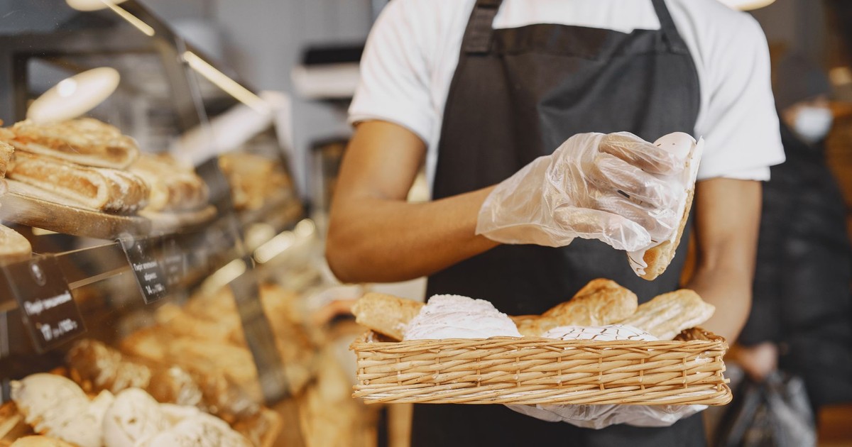 A baker wearing a mask and gloves prepares fresh pastries in a bakery environment.