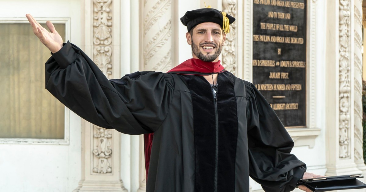 Smiling male graduate in cap and gown celebrating his achievement with arms raised outdoors.