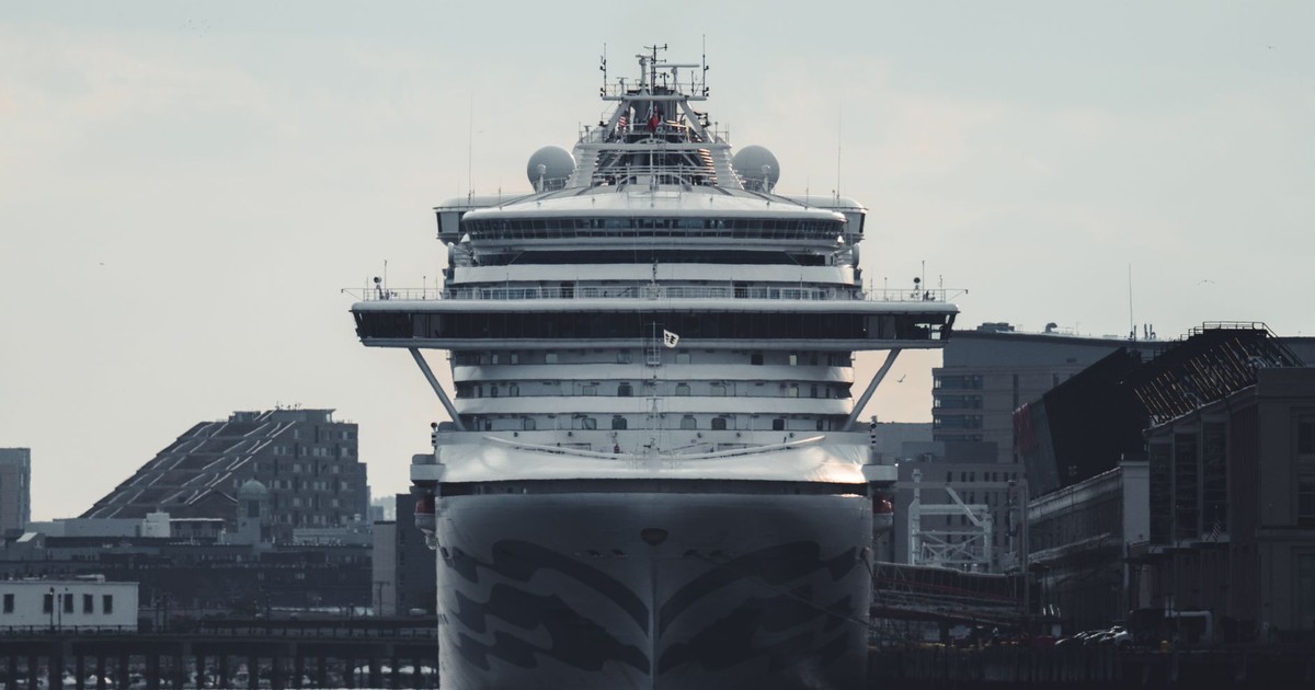 A massive cruise ship docked at an urban harbor, amidst tall buildings and clear skies.
