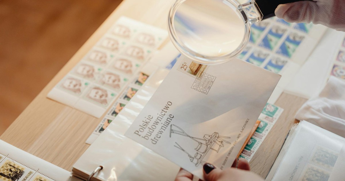 Gloved hands hold a magnifying glass over postage stamps on white paper with a wooden background.