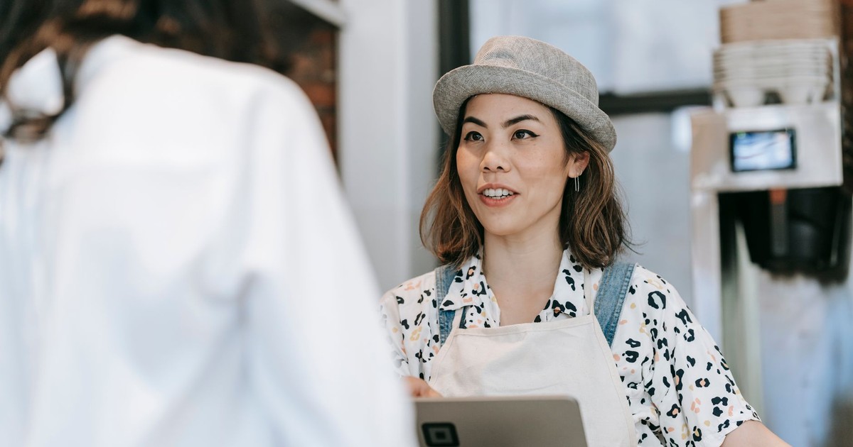 A cheerful barista in a hat takes an order from a customer at a cozy coffee shop counter.