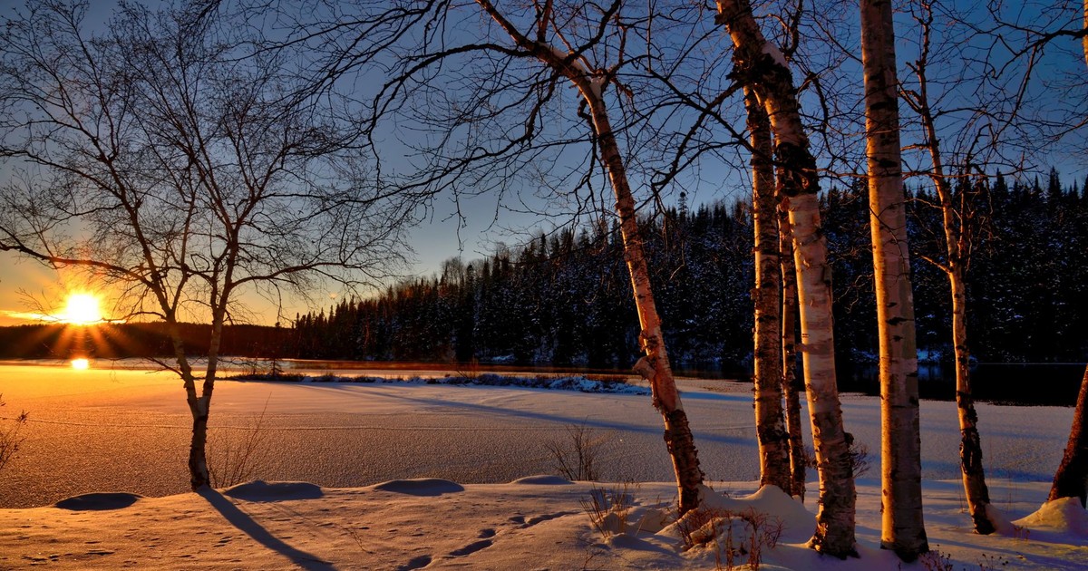 A serene winter sunset casting shadows on a frozen lake surrounded by snow and trees.