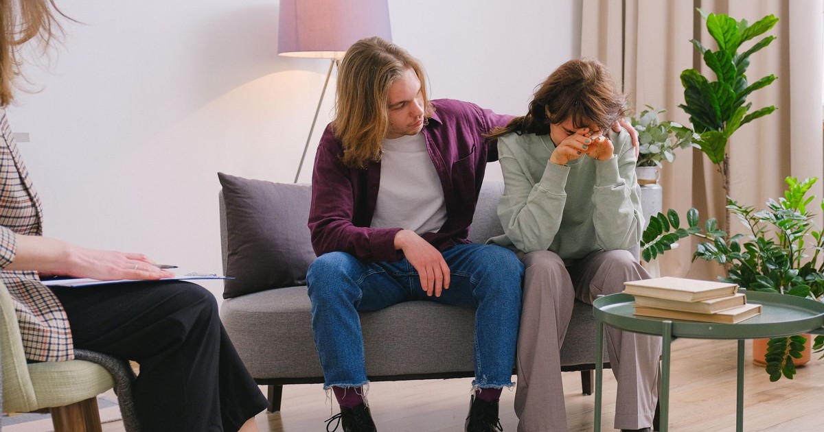 Young male embracing frustrated girlfriend during session with psychologist in light room in daytime