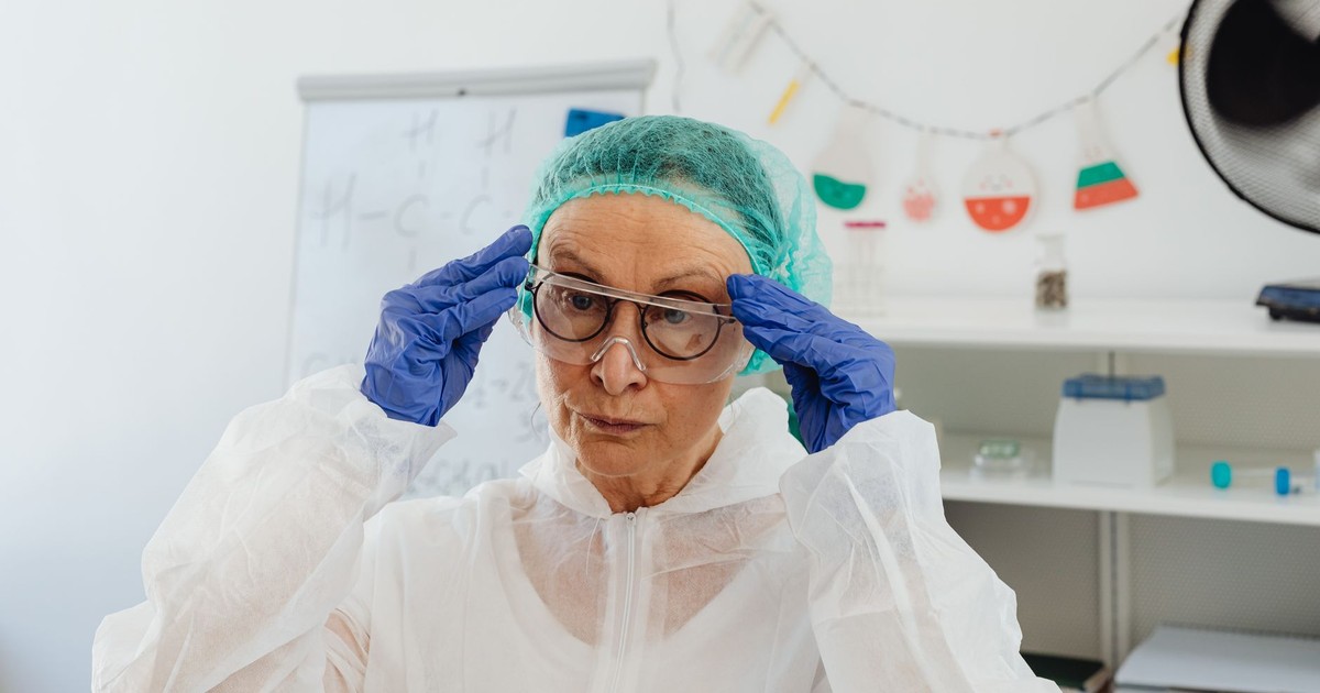 Elderly woman scientist adjusting goggles while wearing full protective lab attire including gloves and gown.