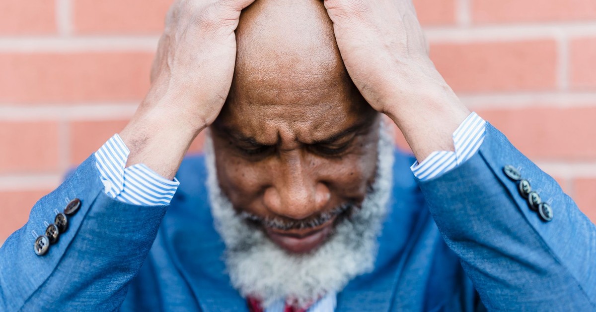 Unhappy bald African American male worker with gray beard in classy wear standing near building with hands on head on street