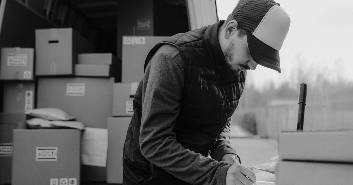 Black-and-white image of a courier documenting package details by a delivery van.