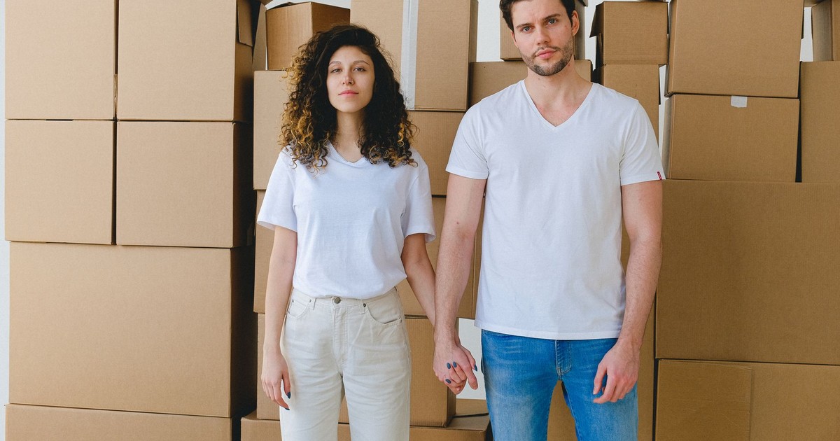 A young couple holding hands in front of a stack of cardboard boxes, possibly indicating moving or relocation.