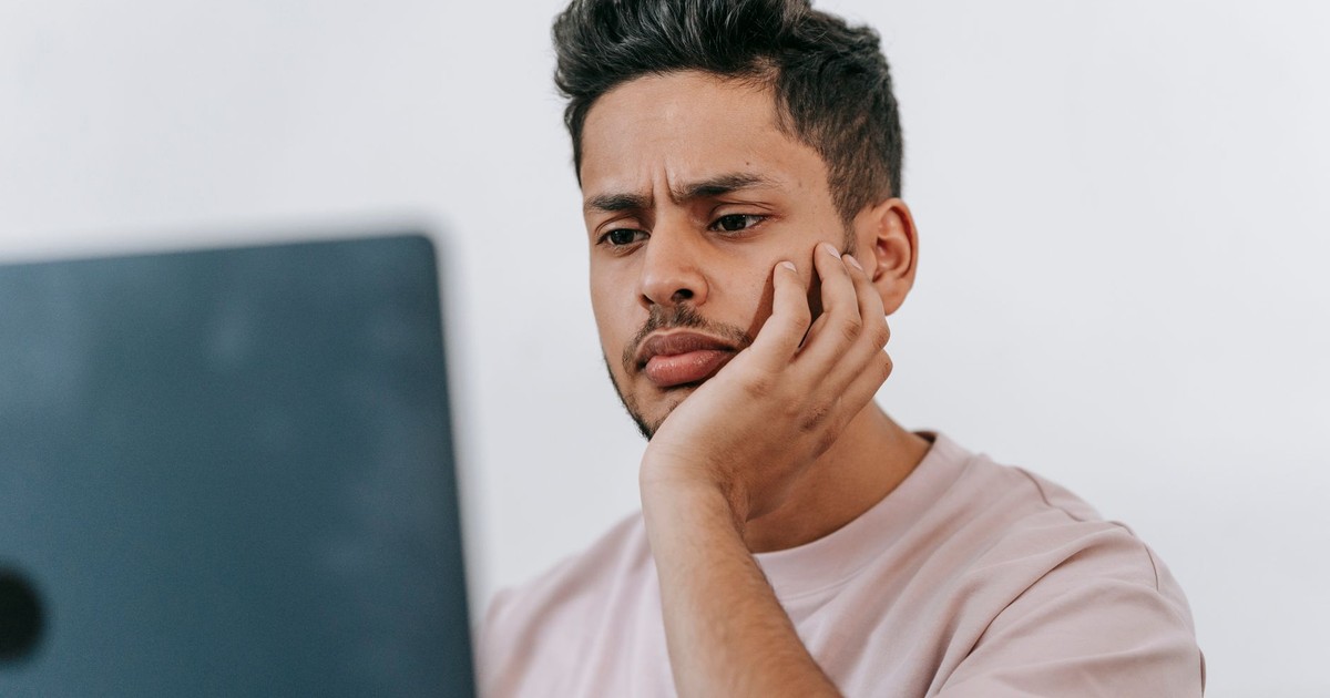 Serious young ethnic male in t shirt browsing netbook and leaning on hand in light room