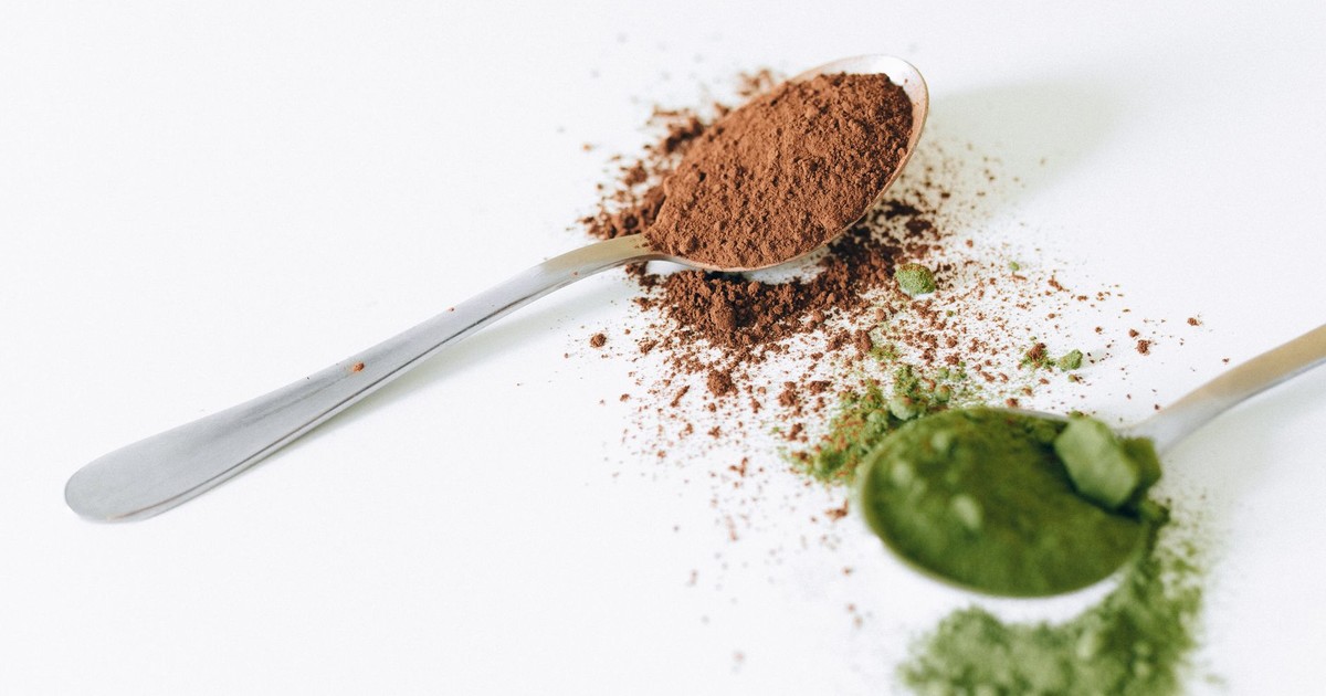 Close-up of cocoa and green tea powder on spoons against a white background.
