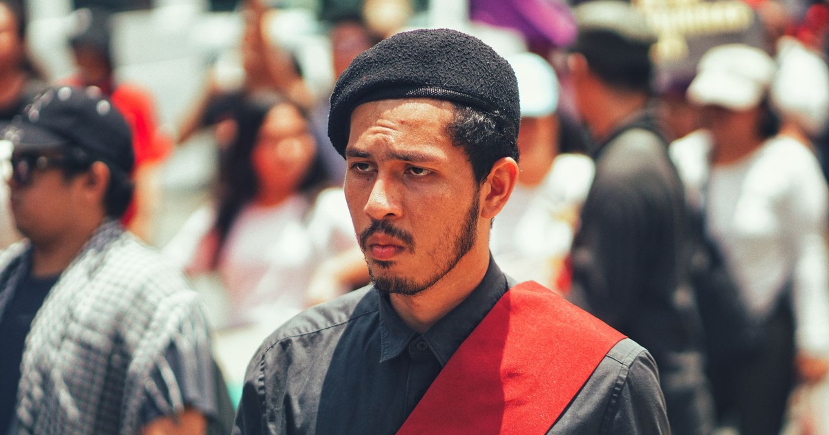 A young man with a red sash and beret focuses intently amidst an outdoor protest crowd.