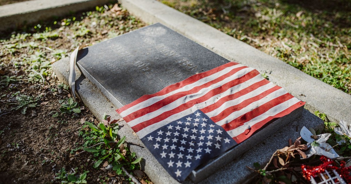 Patriotic tribute with a US flag draped over a tombstone in a serene graveyard setting.