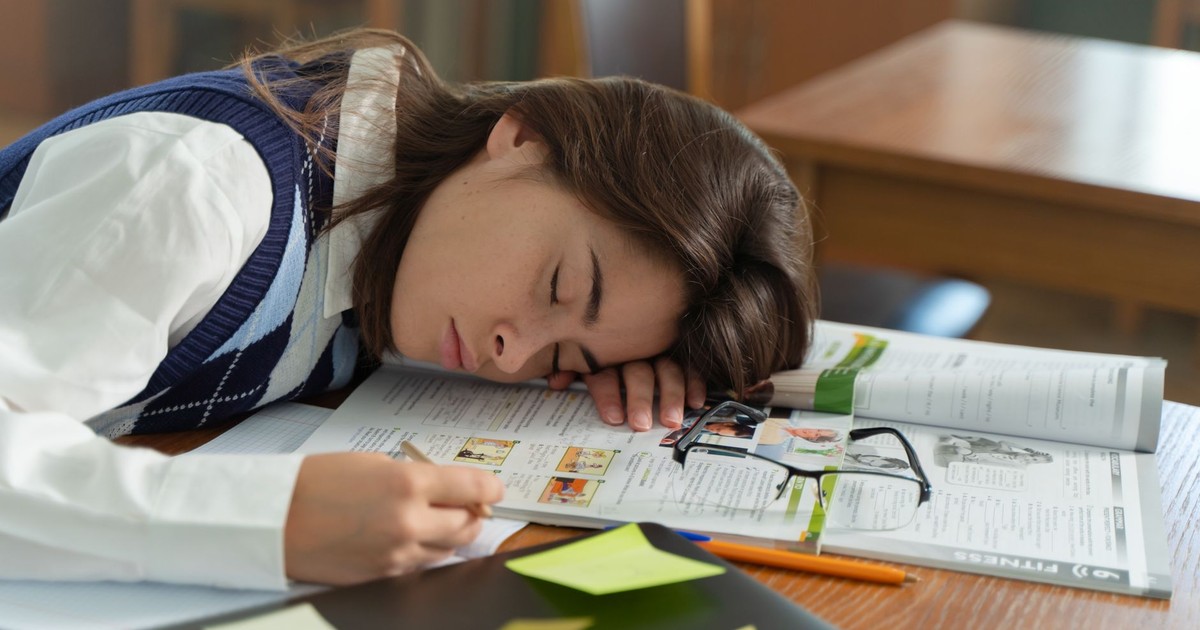A young student sleeps on a school desk, surrounded by open books and study notes.