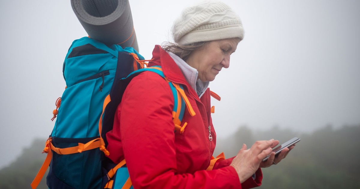 Elderly woman in red jacket checks phone while hiking in foggy scenery outdoors.