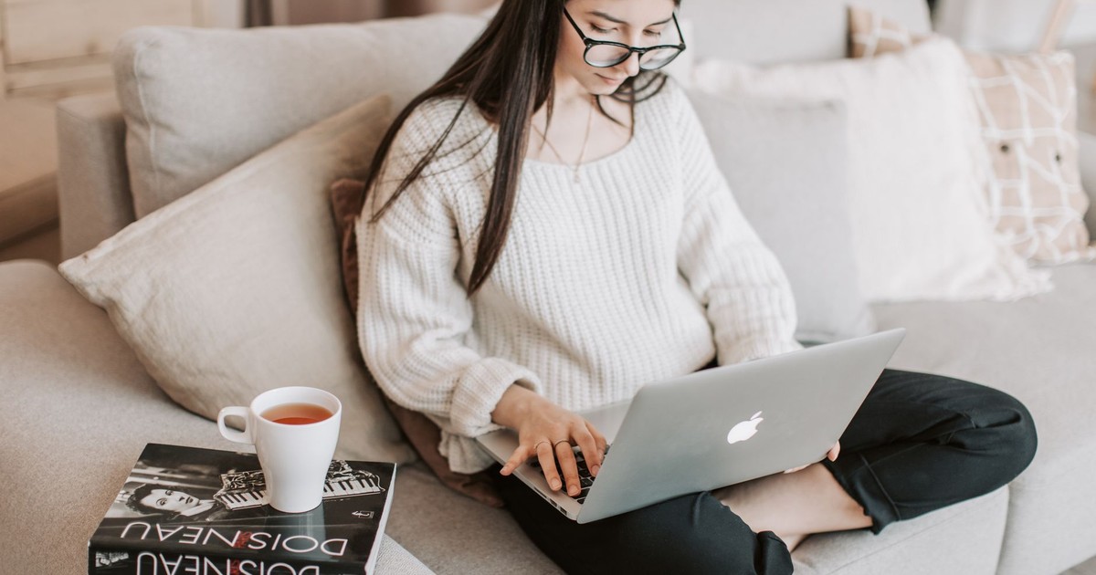 From above crop female in casual clothes and eyeglasses sitting on cozy sofa with book and cup of tea while using laptop for surfing Internet