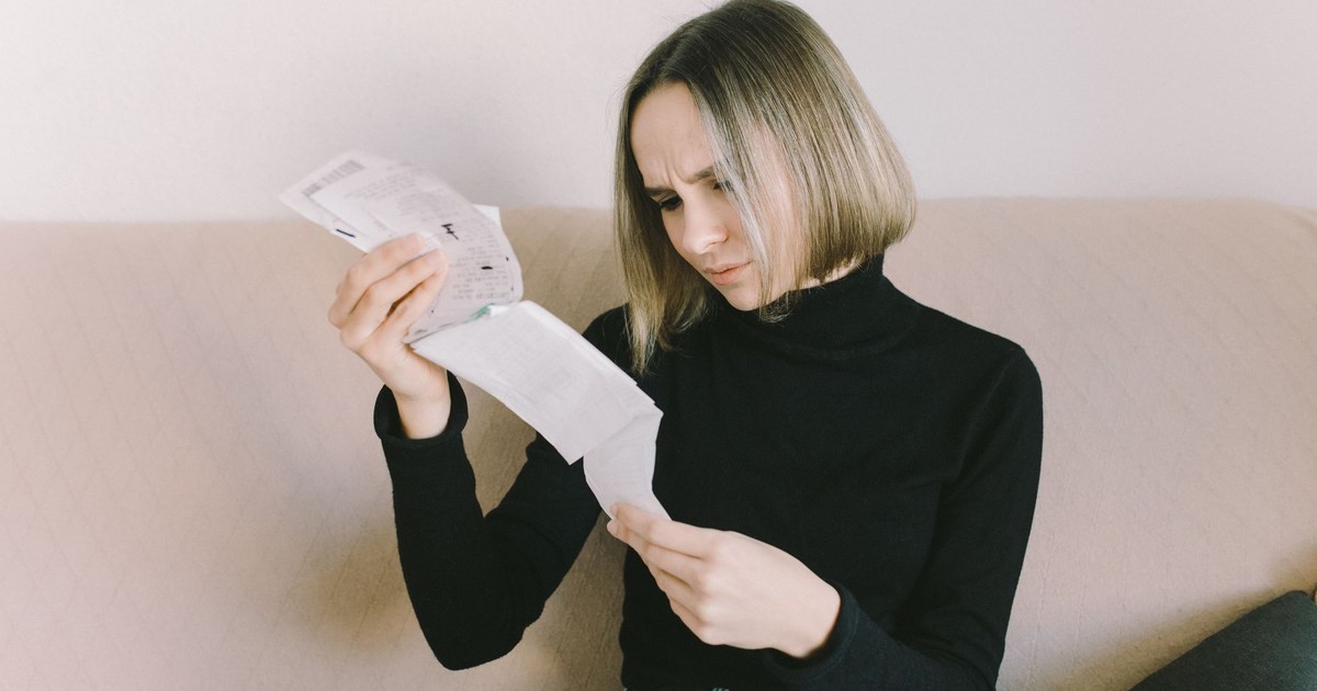Concerned woman in black sweater examining bills on beige sofa indoors.