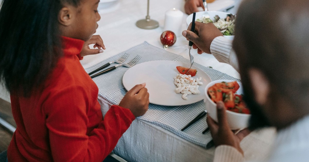 From above of crop anonymous ethnic parent serving pasta salad and tomato slice to girl while celebrating New Year at home