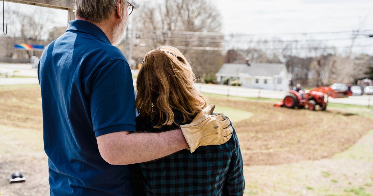 A couple stands arm in arm, observing farmland and a tractor from their porch.