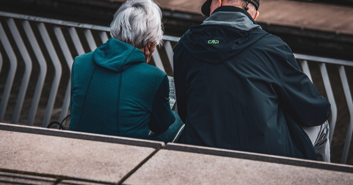 Elderly couple in jackets sitting on steps overlooking an urban setting, captured candidly from behind.