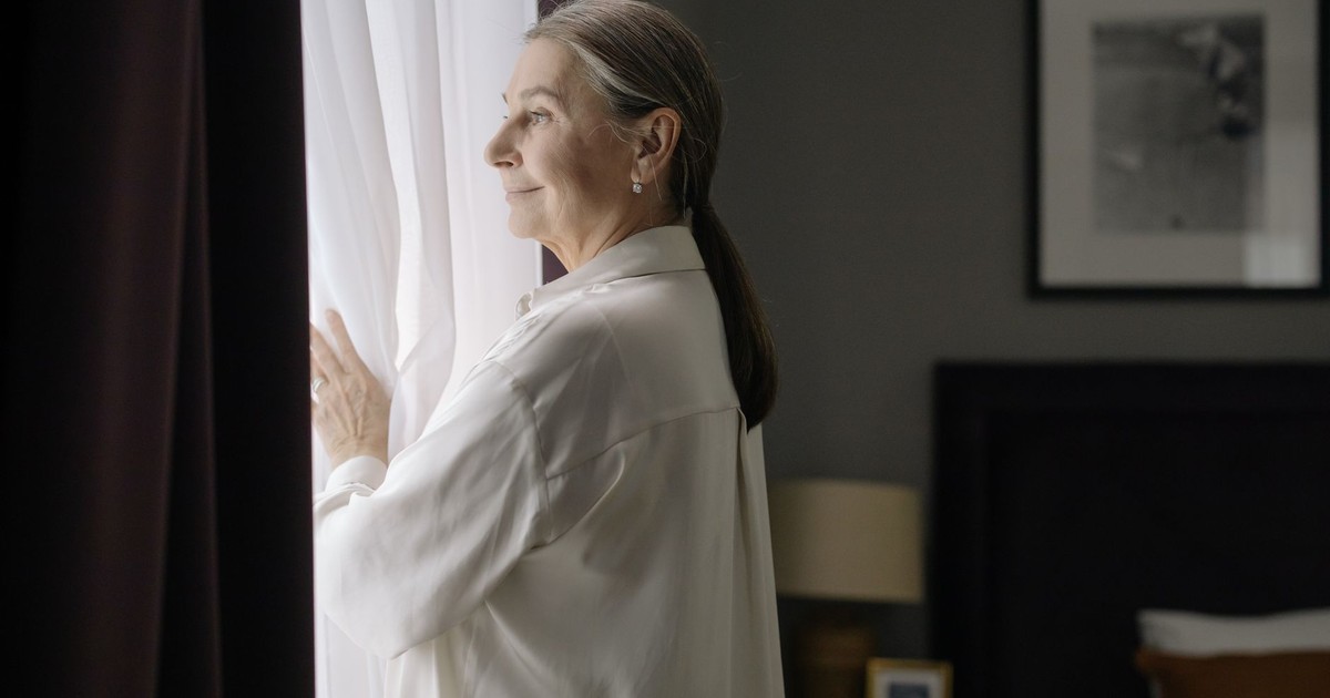 A senior woman with grey hair stands by a window, deep in thought, in a cozy home setting.